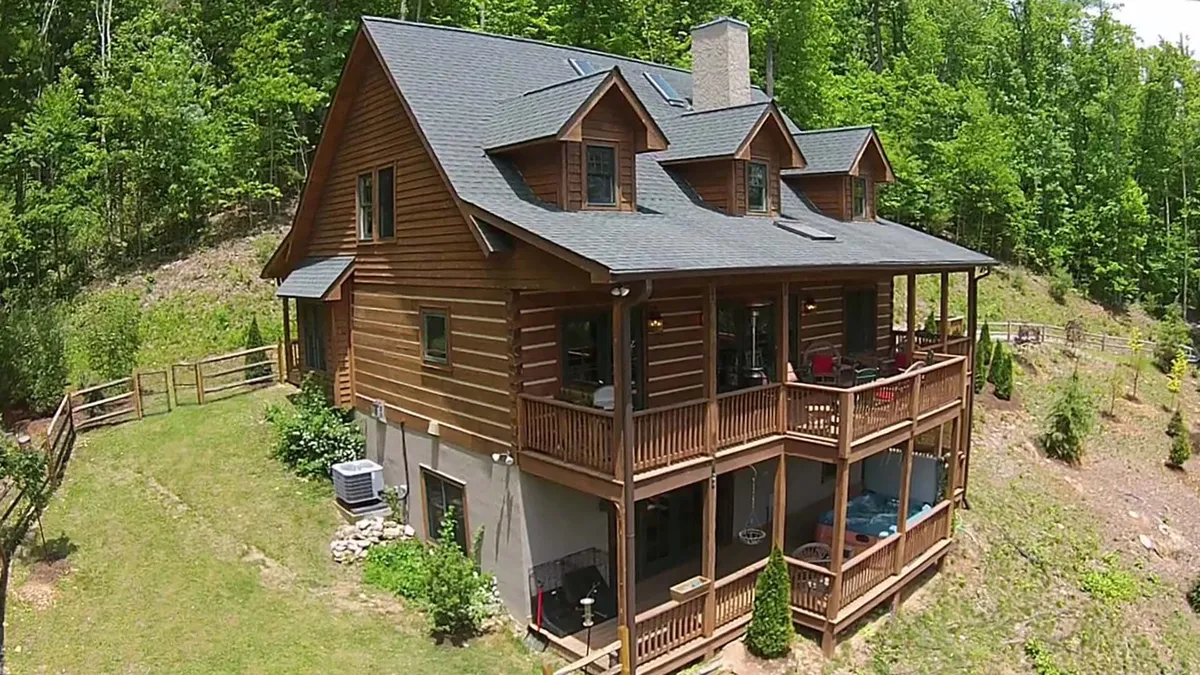 A log home nestled among lush greenery, featuring a spacious porch with seating and ample room for group gatherings. The large wooden structure is surrounded by trees and gives off a relaxed, welcoming atmosphere ideal for outdoor socializing.