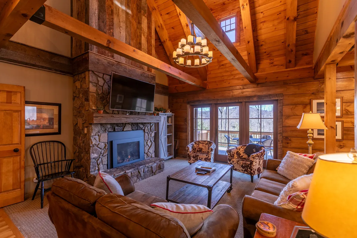 This image showcases a spacious great room with a massive stone fireplace as the focal point, flanked by large windows that let in natural light. The room features wooden beams overhead, plush sofas, and decorative armchairs arranged for conversation or relaxing by the fire. The scene conveys warmth and a welcoming atmosphere, inviting guests to gather and unwind.