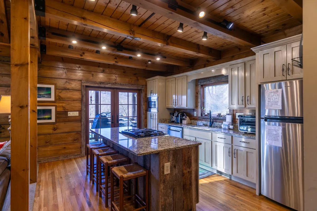 A view of the kitchen area with granite countertops, rustic wooden beams, and stools positioned at the island. A window in the background provides a glimpse of the outdoors, suggesting a seamless flow between indoor comfort and natural beauty.