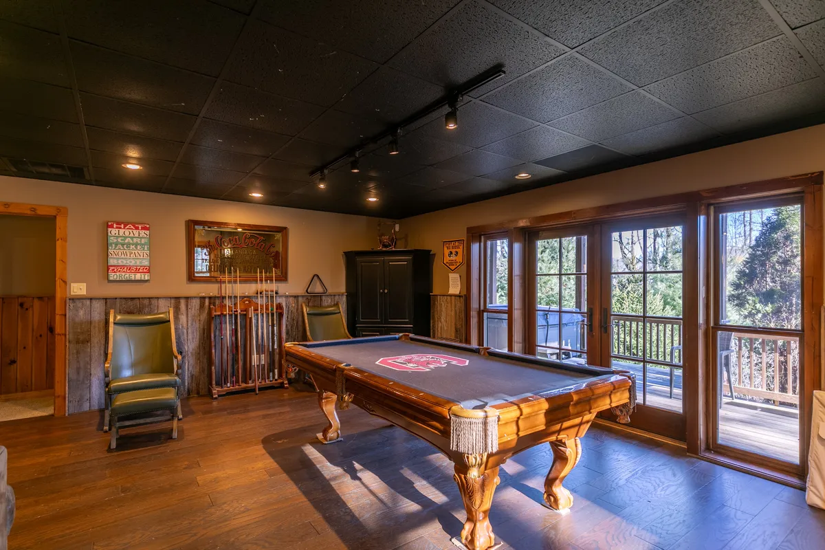 A bright, welcoming recreation room featuring a pool table, large windows with views of the outdoors, wooden accents, and wall art. The space is designed for socializing, relaxing, and enjoying downtime after a day of outdoor activities.