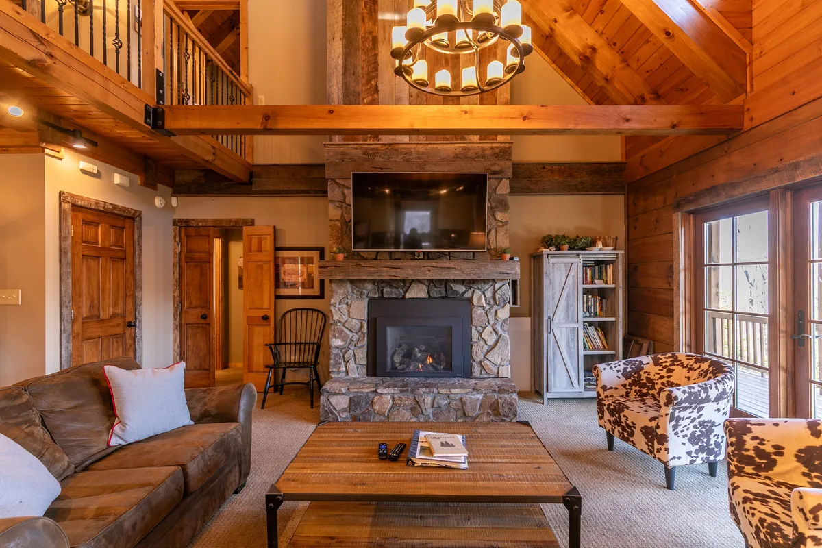 A spacious living area with a grand stone fireplace, comfortable seating, large chandelier, and rustic wooden beams. Through the windows, you can catch glimpses of the mountain landscape outside, evoking a cozy retreat perfect for watching snowfall and relaxing after outdoor adventures.