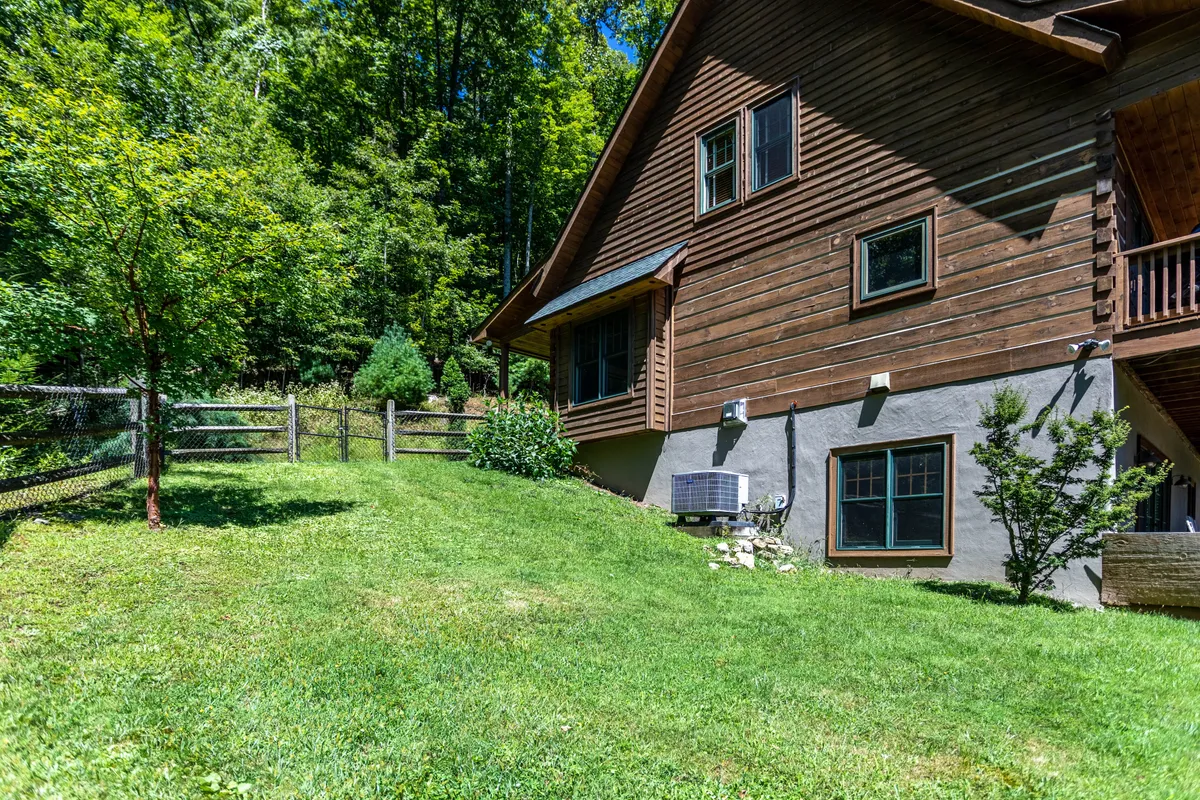A wide view of the home's wooden exterior, showcasing a spacious green lawn surrounded by mature trees and a fenced yard. The scene suggests ample space for outdoor play, family gatherings, and relaxation in nature right outside the front door.