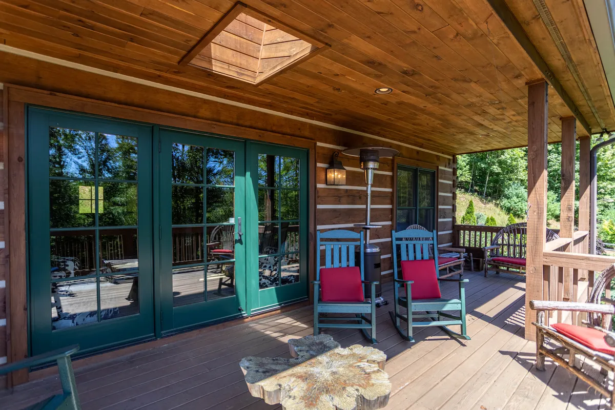 The natural wood deck is furnished with green double doors, wooden rocking chairs, and a stone coffee table. The porch looks out toward the trees, creating a perfect spot for relaxing together, whether with morning coffee or sunset drinks, surrounded by the sights and sounds of nature.