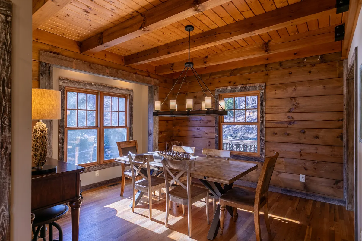 A wooden dining table sits beneath a stylish chandelier, surrounded by large windows that offer picturesque views. The dining area is spacious enough for a group, radiating a sense of togetherness and celebration. The natural light and wooden accents enhance the cozy, communal vibe.