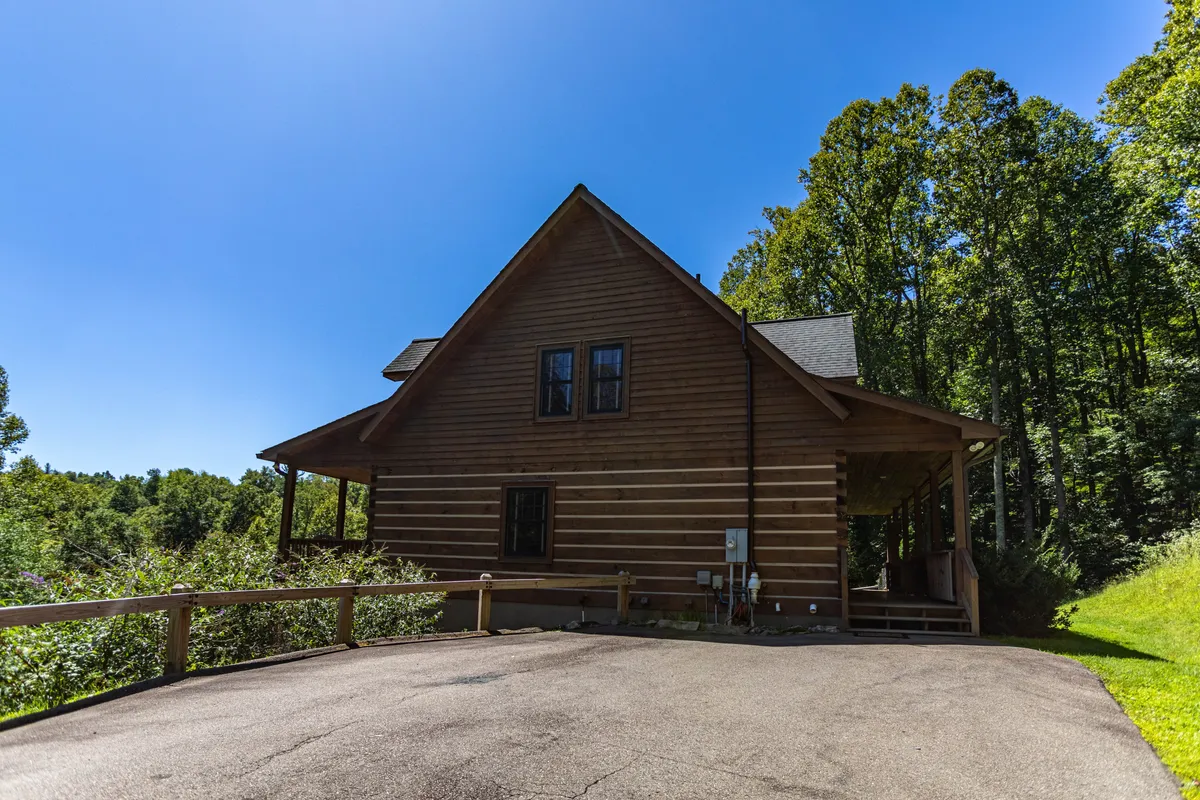 A wider exterior perspective of the property shows classic log cabin architecture set deep within the surrounding forest, with a driveway leading to the home. The tranquil, wooded scenery and inviting porch highlight the sense of seclusion and connection with nature that make the setting ideal for a couple’s retreat.