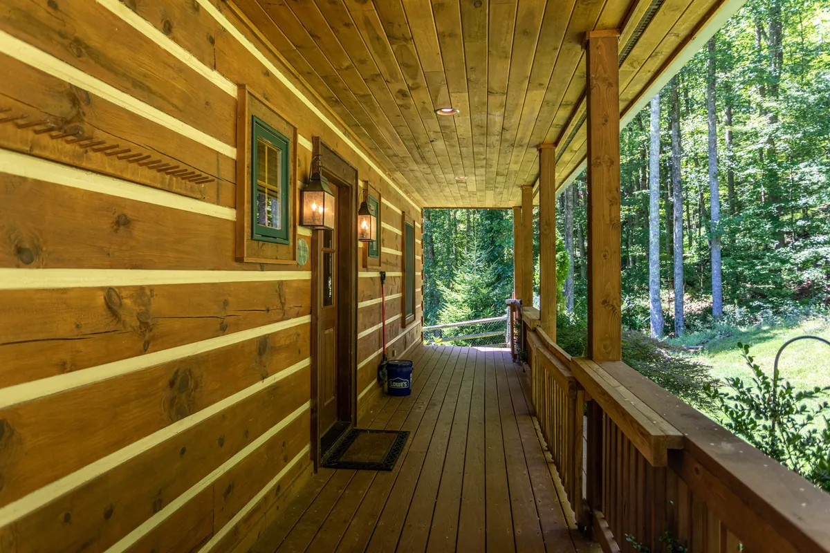 A welcoming exterior view of the property, featuring a wooden door framed by rustic wooden siding, soft light fixtures by the entrance, and green foliage that hints at the cabin’s natural mountain setting. The inviting porch offers a glimpse into the cozy ambiance awaiting inside.