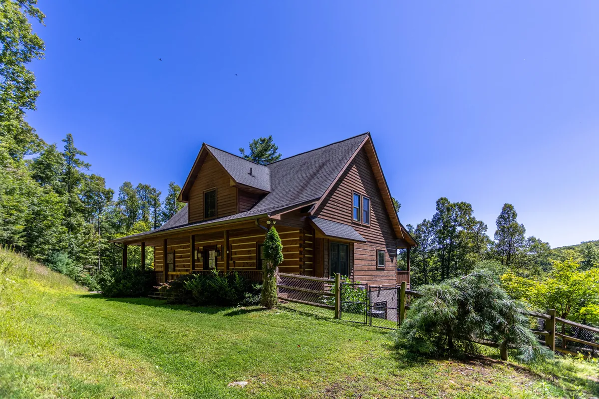 The image showcases an inviting log cabin nestled amidst lush greenery, with a fenced yard suggesting privacy and safety. The spacious covered porch offers views ideal for sunset watching and stargazing, while the natural setting evokes a sense of calm and escape.