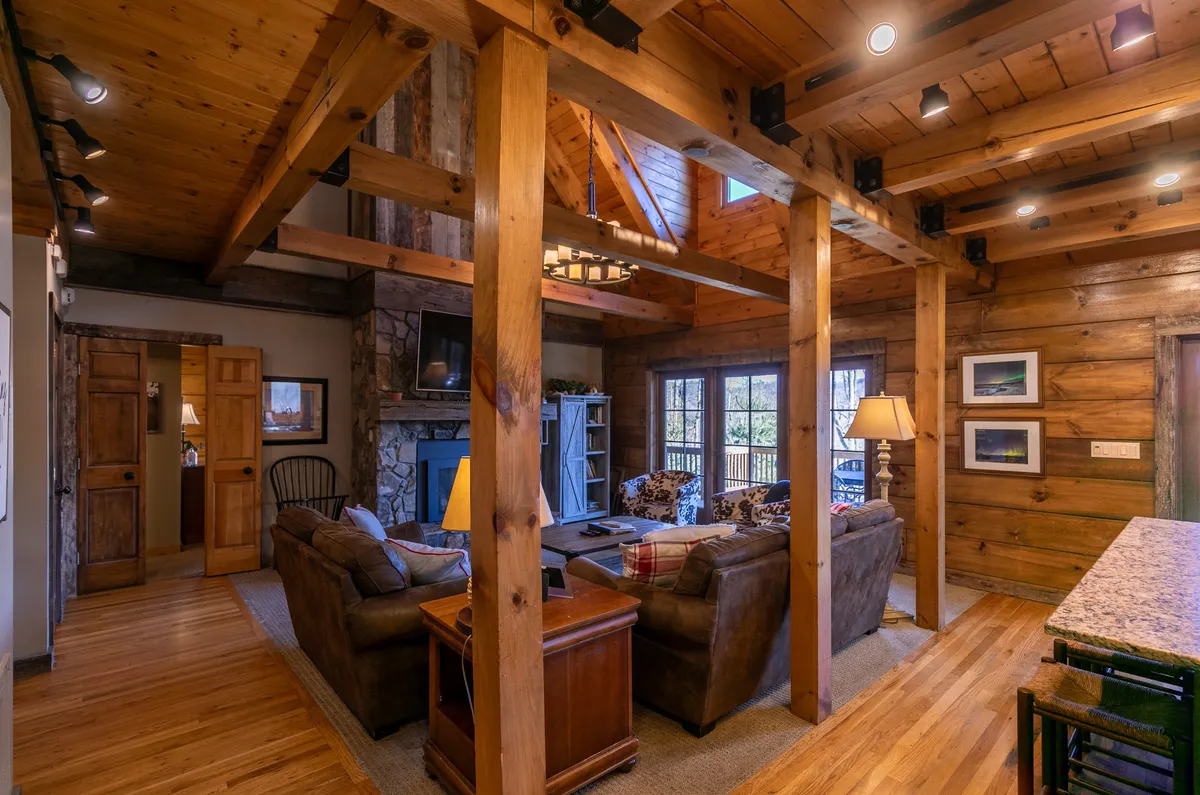 A spacious great room with exposed wooden beams, a welcoming stone fireplace, and a comfortable seating area. Large windows bathe the room in natural light and offer views of the surrounding landscapes, underscoring the seasonal connection between indoors and outdoors.
